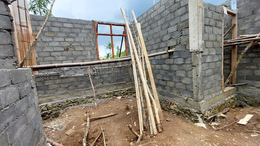 Ein halbfertiges Zimmer ohne Dach mit unverputzten Wänden aus grauen Mauersteinen, an den Ecken ragt Baustahl heraus, die Fenster- und Türrahmen aus Holz sind leer, es liegen Holzlatten und Bambusstangen herum, der Boden besteht aus unebener festgetretener Erde.