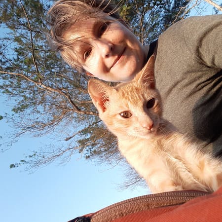 Auf dem Selfie lächelt Steffi in die Kamera. Neben Steffi sitzt ein orange getigertes Kitten auf einem braunroten Rucksack (der ist nur angeschnitten zu sehen) und schaut neugierig auf das Telefon, mit dem das Selfie gemacht wurde. Im Hintergrund sind blauer Himmel und Nadelbäume sichtbar.