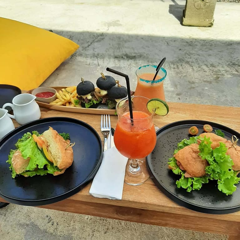 Auf einem niedrigen Holztisch stehen zwei schwarze Teller mit Sandwiches darauf, im Hintergrund steht ein länglicher schmaler Holzteller mit drei schwarzen Miniburgern und Pommes, dazwischen liegt Besteck und stehen Gläser mit Säften.